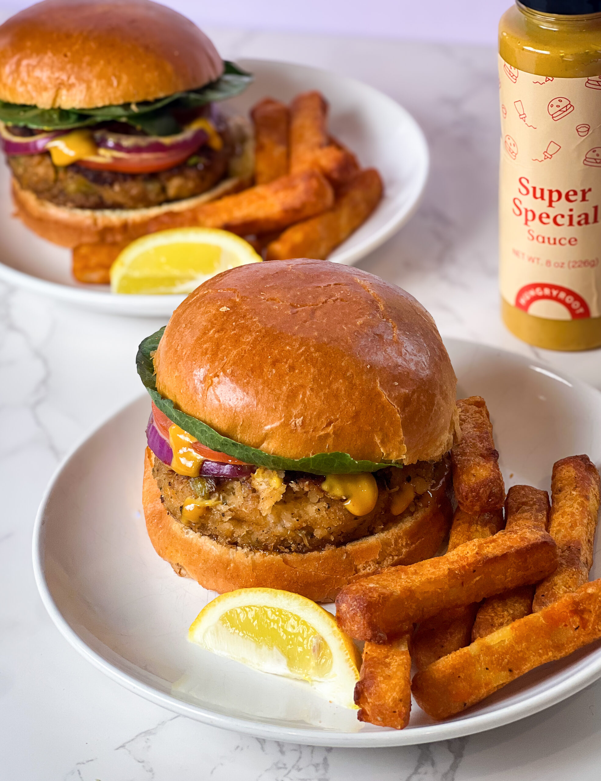 Two plates of mouthwatering crab sandwiches with thick golden fries are shown. Each burger features lettuce, tomato, onion, and sauce in a bun, complemented by a wedge of lemon and a bottle of Super Special sauce on a sleek white marble surface.