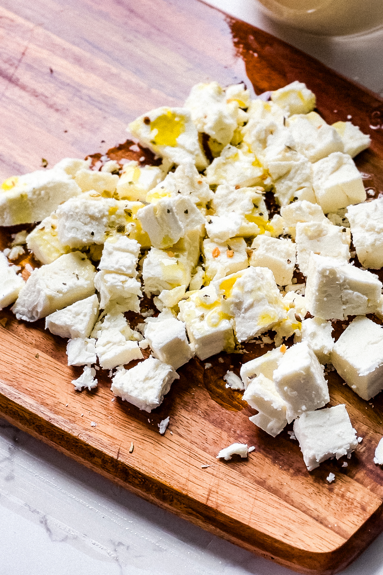 Chopped pieces of white feta cheese, sprinkled with seasoning and drizzled with oil, are spread on a wooden cutting board beside a vibrant quinoa salad with green olives. The surface is lightly stained with oil.