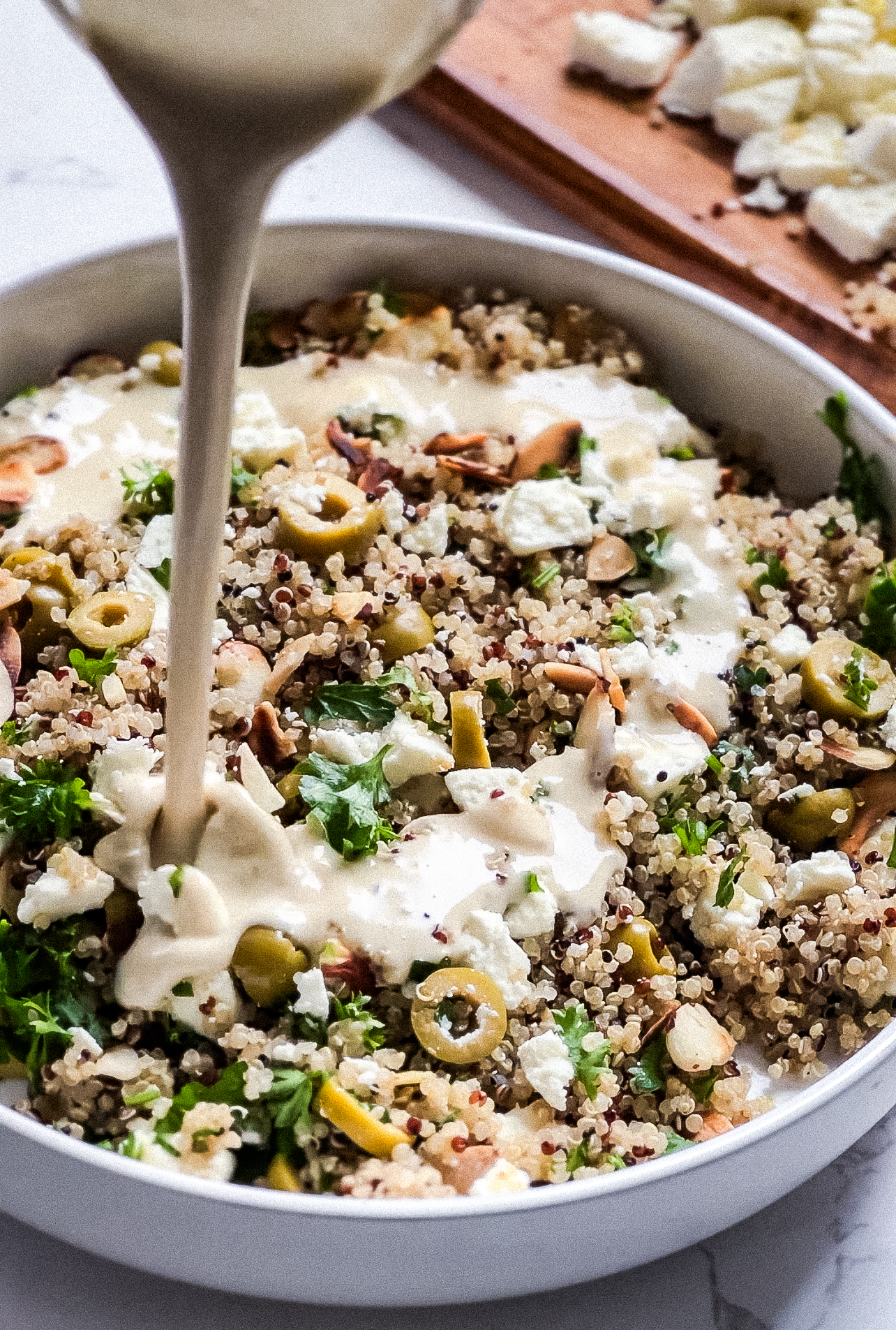 A bowl of crunchy quinoa salad with chopped parsley, green olives, almonds, and crumbled feta is being drizzled with dressing from above. A wooden board with more feta is in the background.