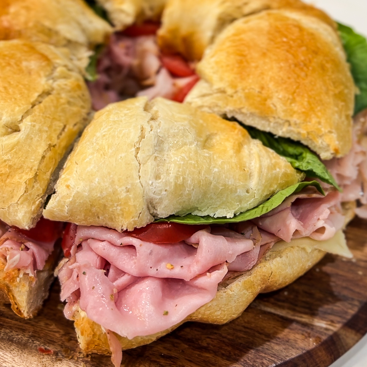 A sliced sandwich ring with layers of ham, cheese, lettuce, and tomatoes on a wooden board. The bread is lightly toasted and golden brown.