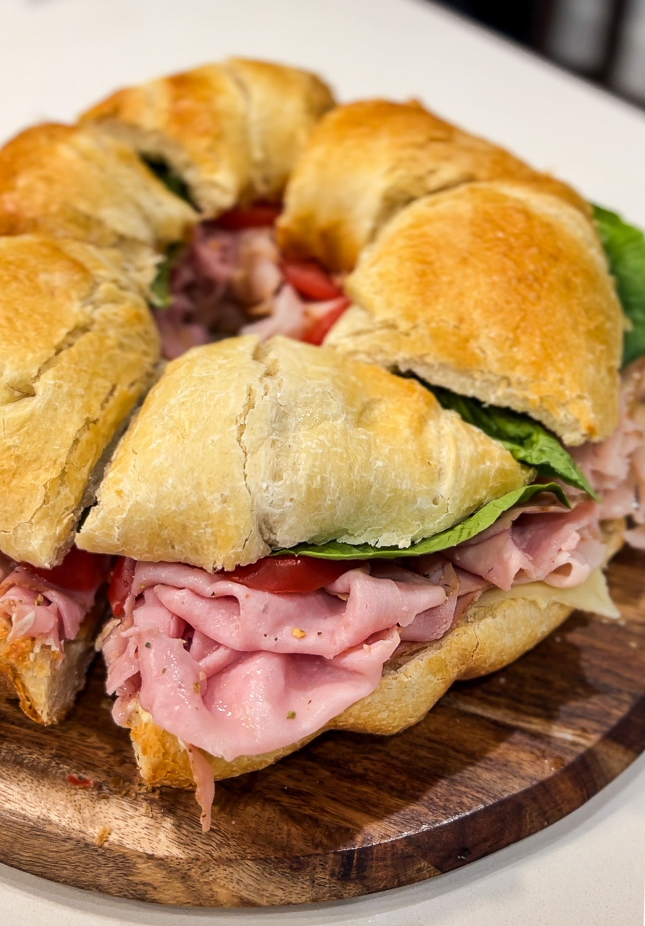 A large sandwich on a wooden board, made with a round loaf of crusty bread. It is filled with layers of sliced ham, lettuce, and tomatoes. The sandwich is cut into wedges, revealing the ingredients inside.