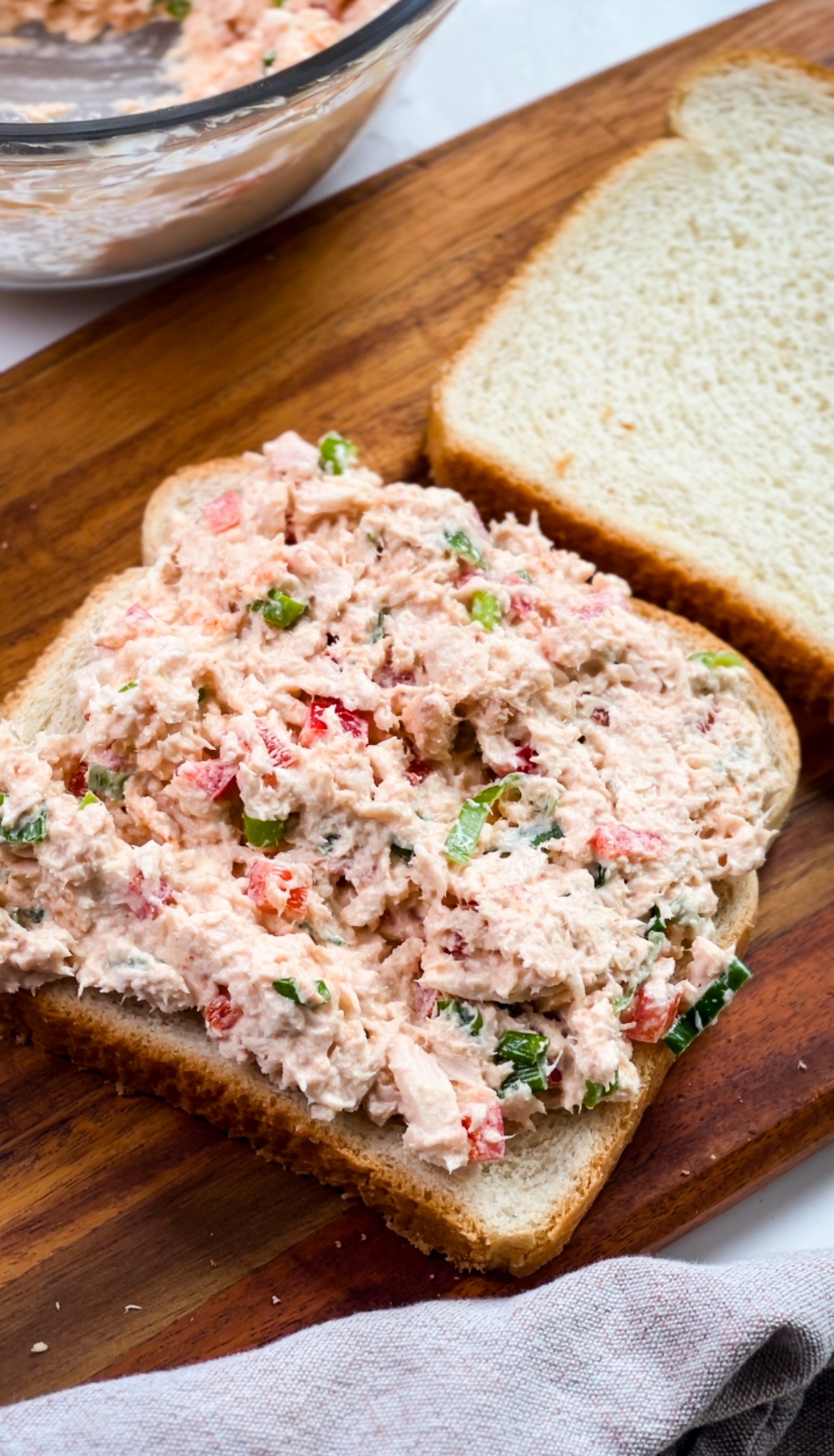 A sandwich being prepared with a generous spread of tuna salad on a slice of white bread, resting on a wooden cutting board. The salad is mixed with diced veggies, giving it a colorful appearance. Another slice of bread is nearby, ready to complete the sandwich.