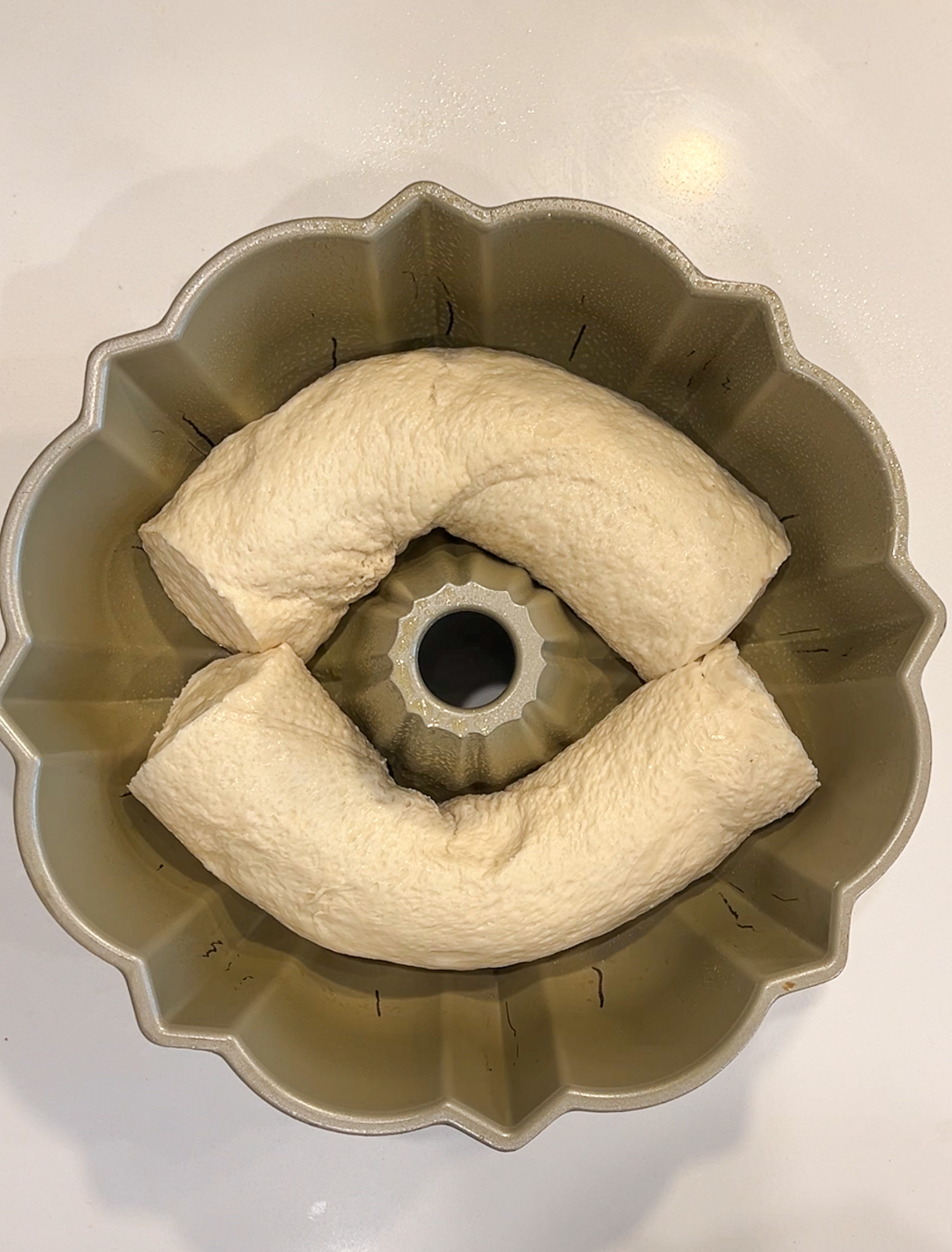 Raw dough shaped into a ring is placed in a Bundt cake pan on a white surface. The dough is slightly overlapping at the edges, forming an irregular circle within the fluted pan.