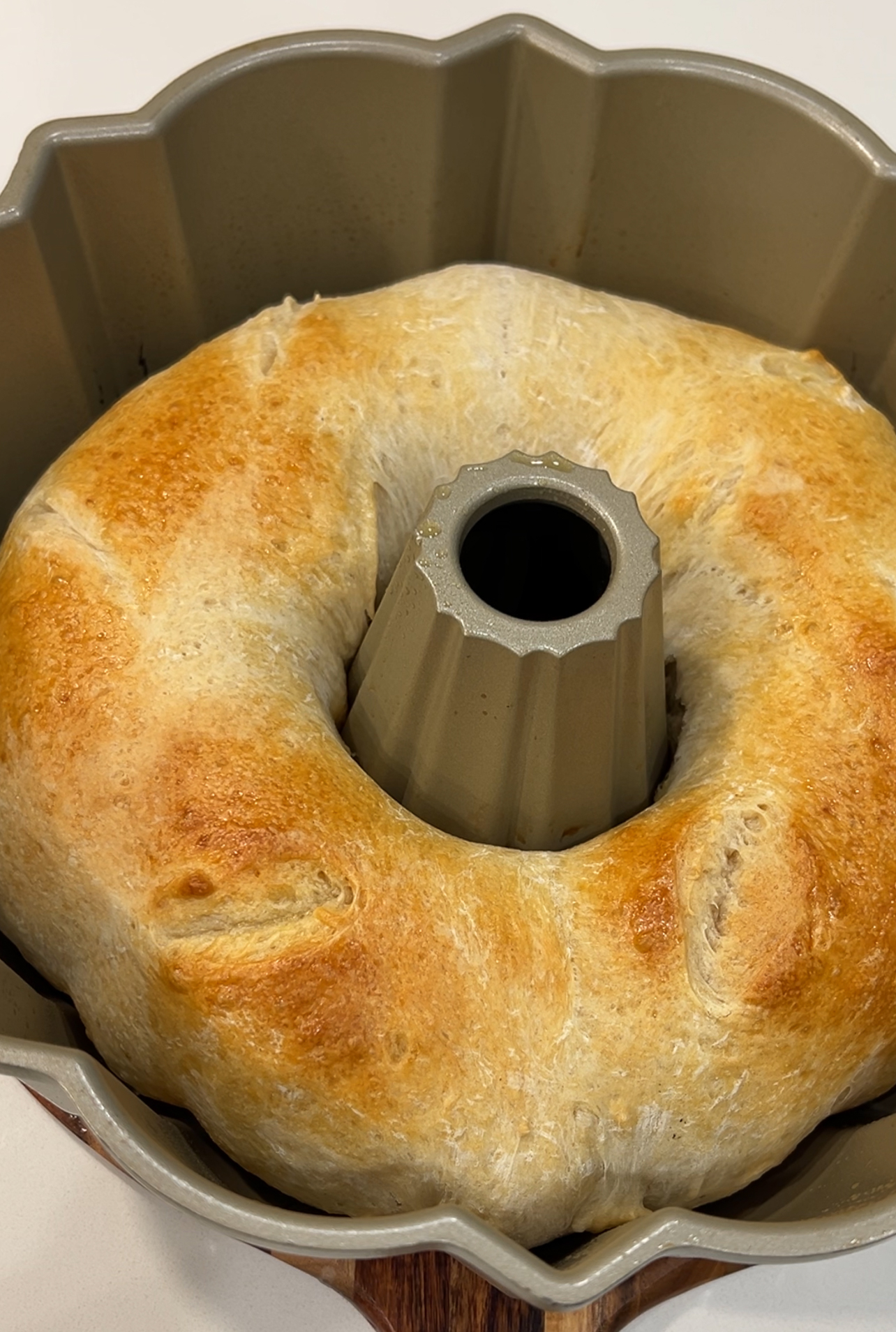 A freshly baked golden-brown bread ring sits in a bundt pan. The bread has a smooth, shiny crust and a soft texture, with slight browning on the surface, creating an appetizing appearance.