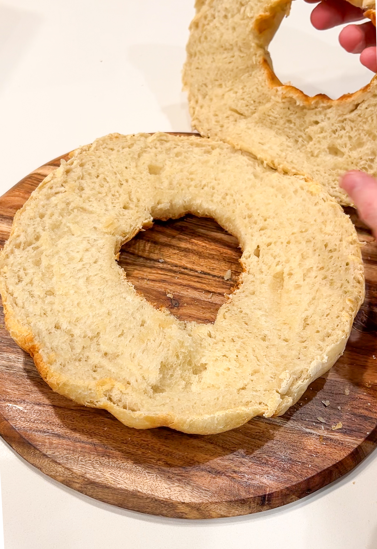 A sliced bagel is placed on a wooden board with one half lying flat and the other half being held slightly above by a hand. The bagel has a golden-brown crust and a soft, fluffy interior.