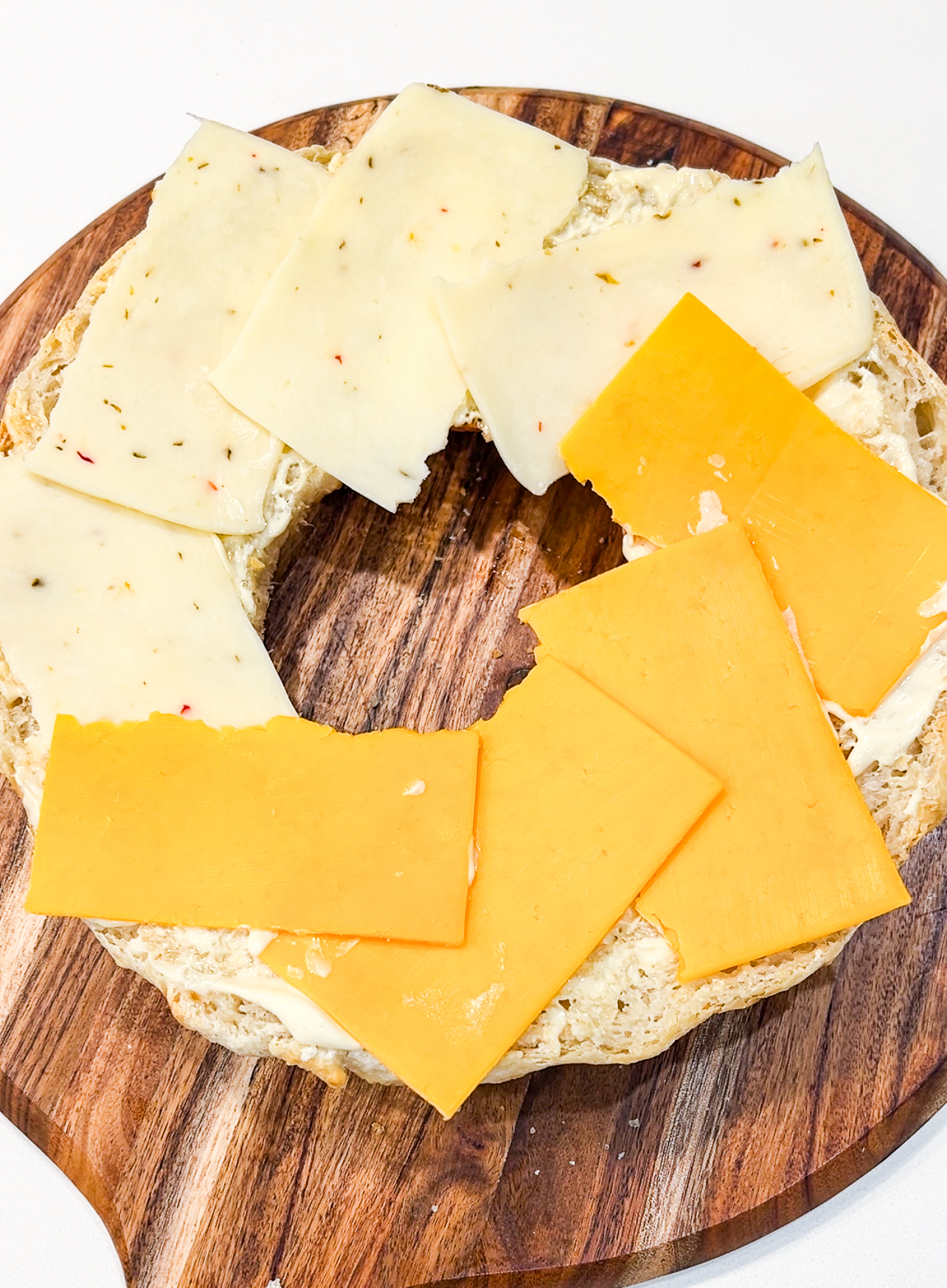 A sliced bagel topped with overlapping slices of white and orange cheddar cheese on a wooden cutting board. The cheese covers most of the surface of the bagel.