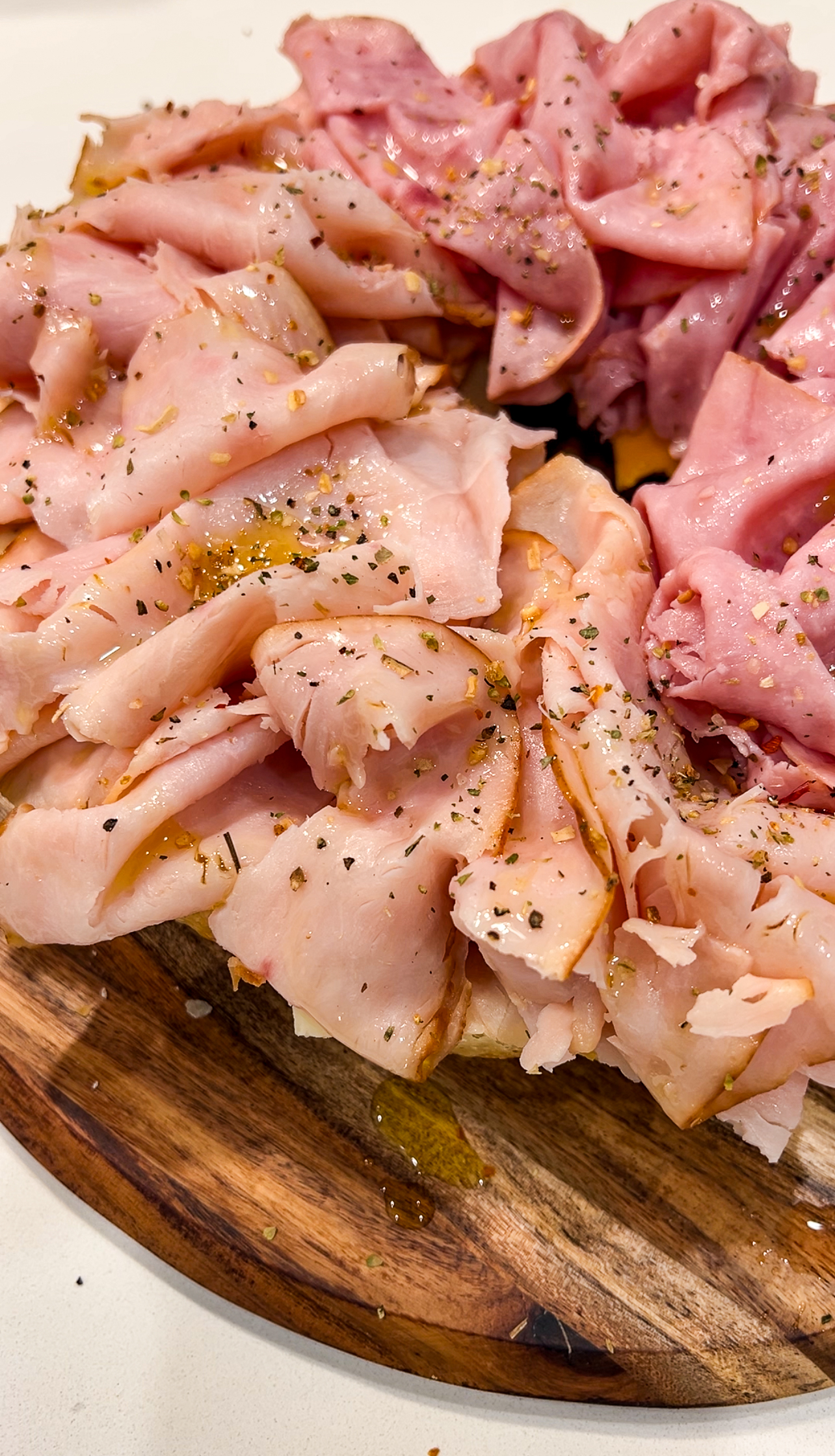 Close-up of thinly sliced roast pork arranged on a wooden board. The meat is garnished with a sprinkle of herbs and drizzled with olive oil, giving it a glistening appearance. The arrangement highlights the meats pink and tender texture.