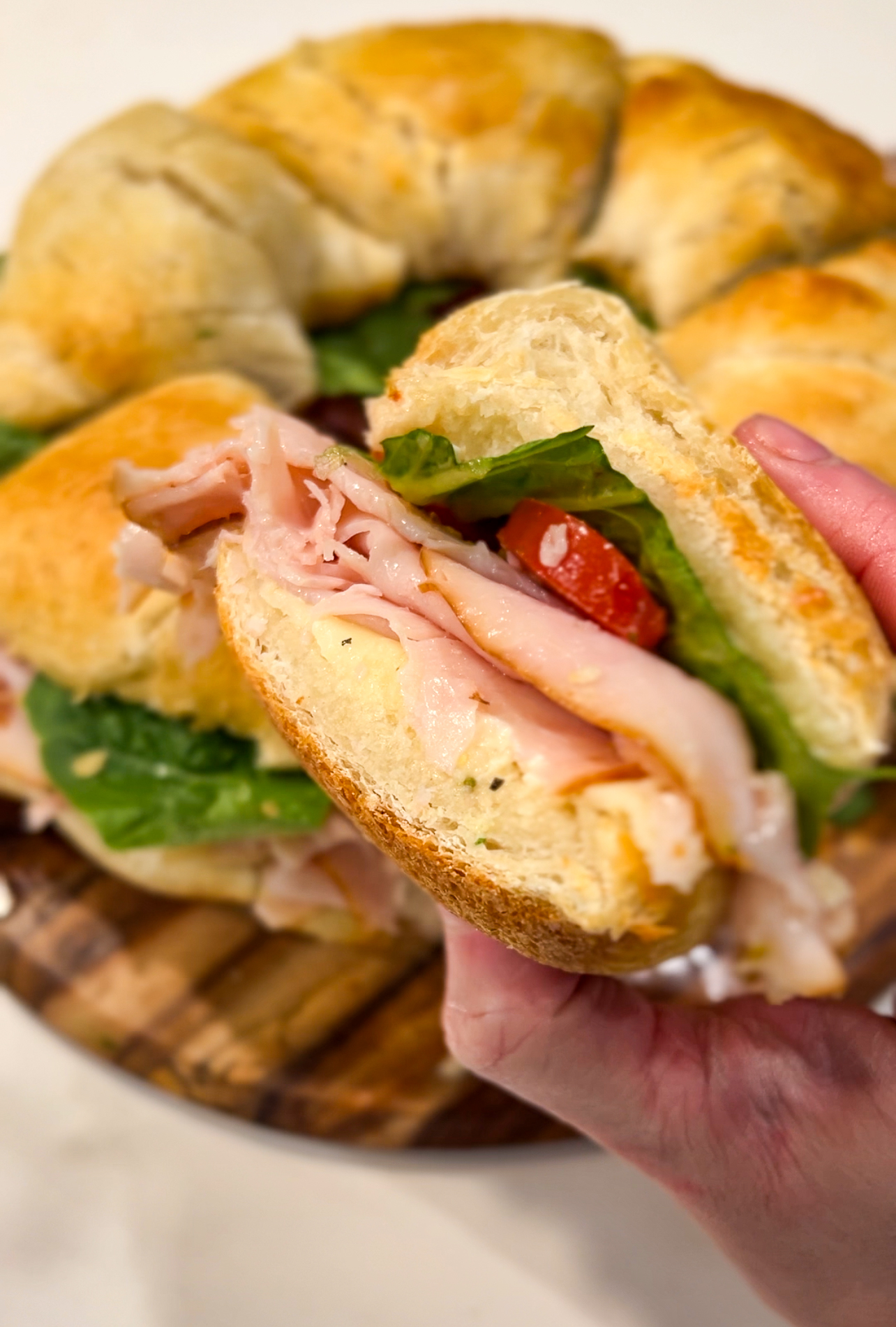 Close-up of a hand holding a sandwich filled with turkey slices, lettuce, and tomato between sliced bread. More sandwiches are visible in the background, arranged on a wooden surface.