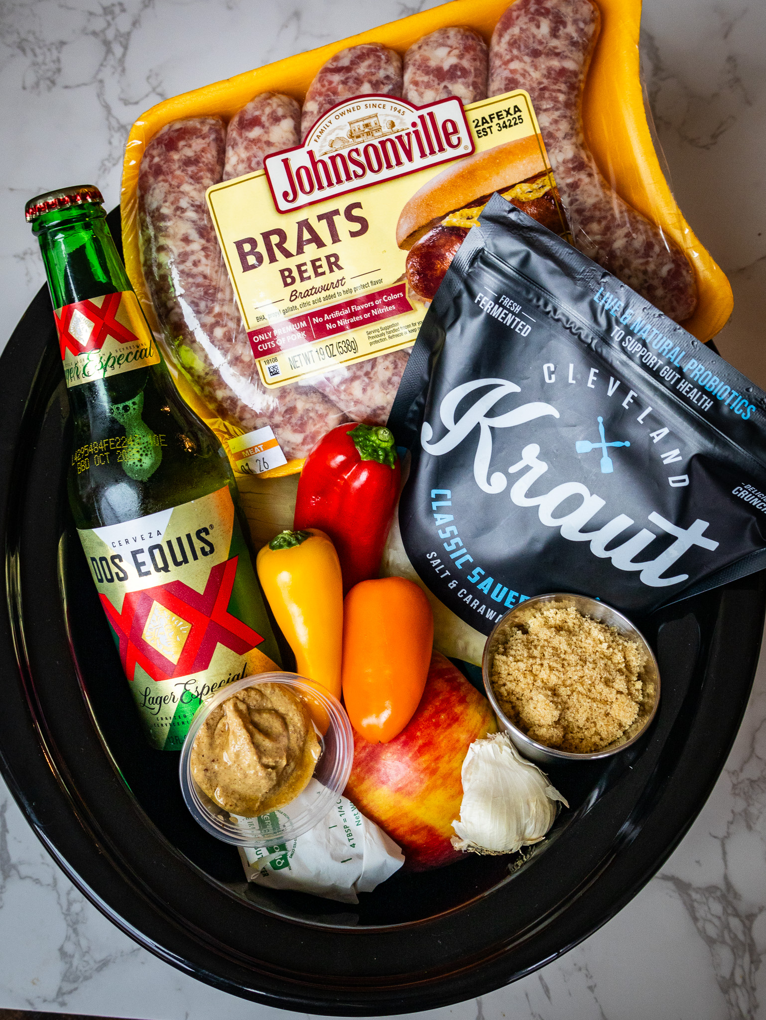 A black tray on a marble surface holds a pack of Johnsonville beer brats, Dos Equis beer, colorful peppers, Cleveland Kraut, an apple, a garlic bulb, a small container of mustard, and a container of brown sugar.