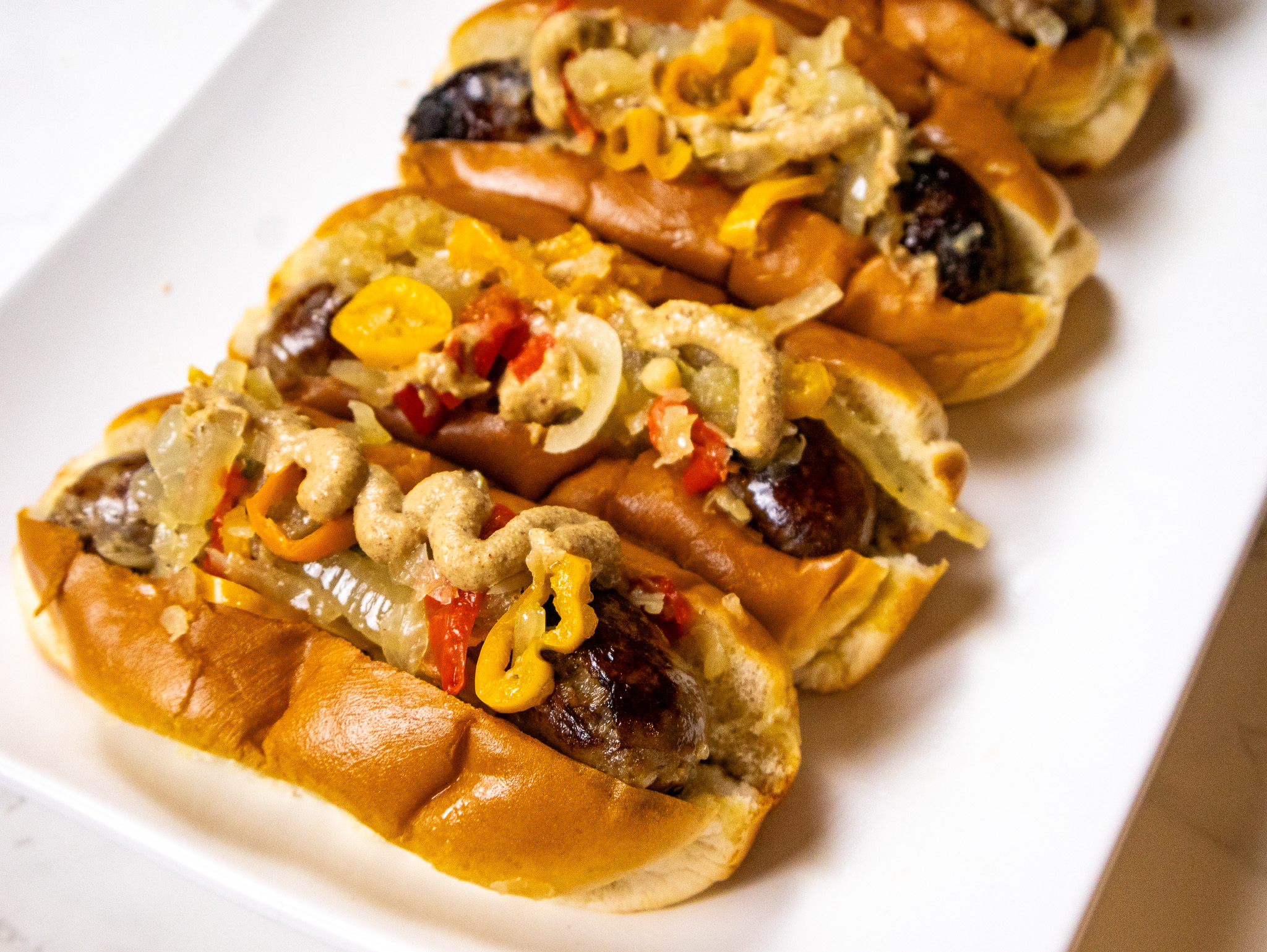 Close-up of three grilled sausages in buns, topped with sautéed onions, mixed bell peppers, and mustard. The sausages are arranged in a row on a white plate.