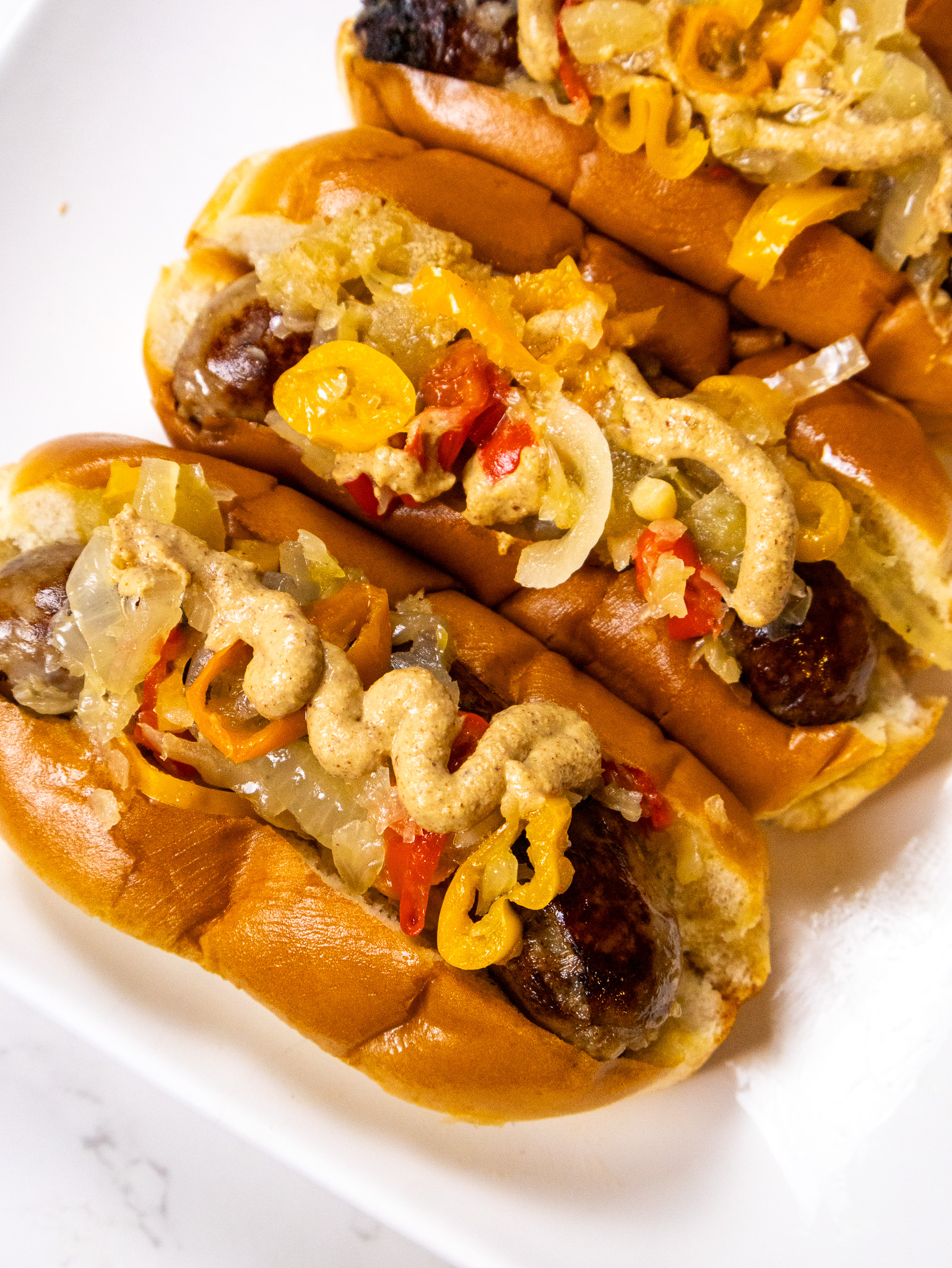 Close-up of three grilled sausages in buns, topped with sautéed onions, yellow peppers, and a thick drizzle of mustard. They are arranged on a white plate, highlighting the vibrant colors and textures of the toppings.