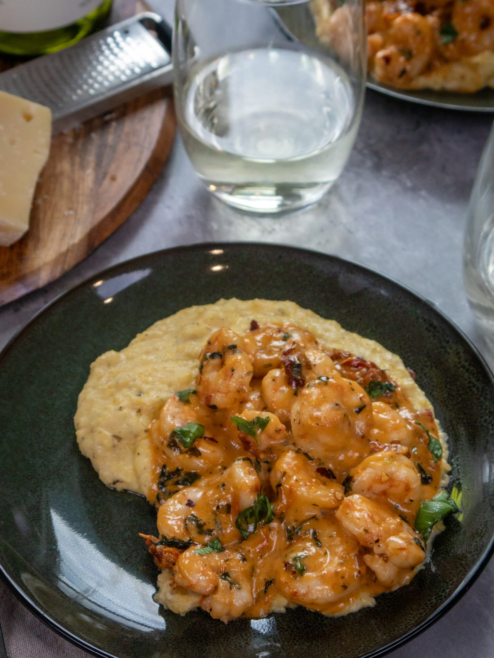 A plate of creamy Parmesan polenta topped with seasoned shrimp in a marry me sauce, garnished with herbs. A glass of white wine sits beside the dish, and a board with cheese and a grater is in the background—perfect for a romantic Valentines Day dinner.