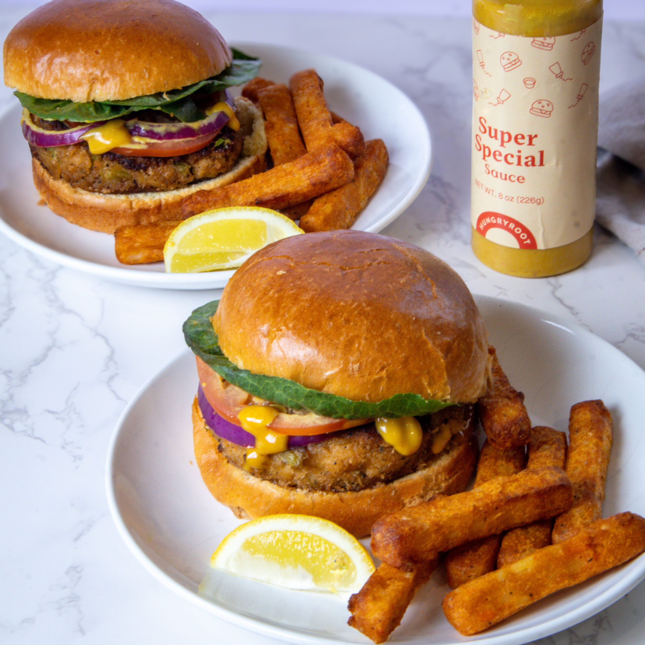 Two plates with veggie burgers on brioche buns, topped with lettuce, onion, tomato, and cheese, served with thick-cut fries and lemon wedges. A bottle of Super Special Sauce stands nearby on a marble surface.