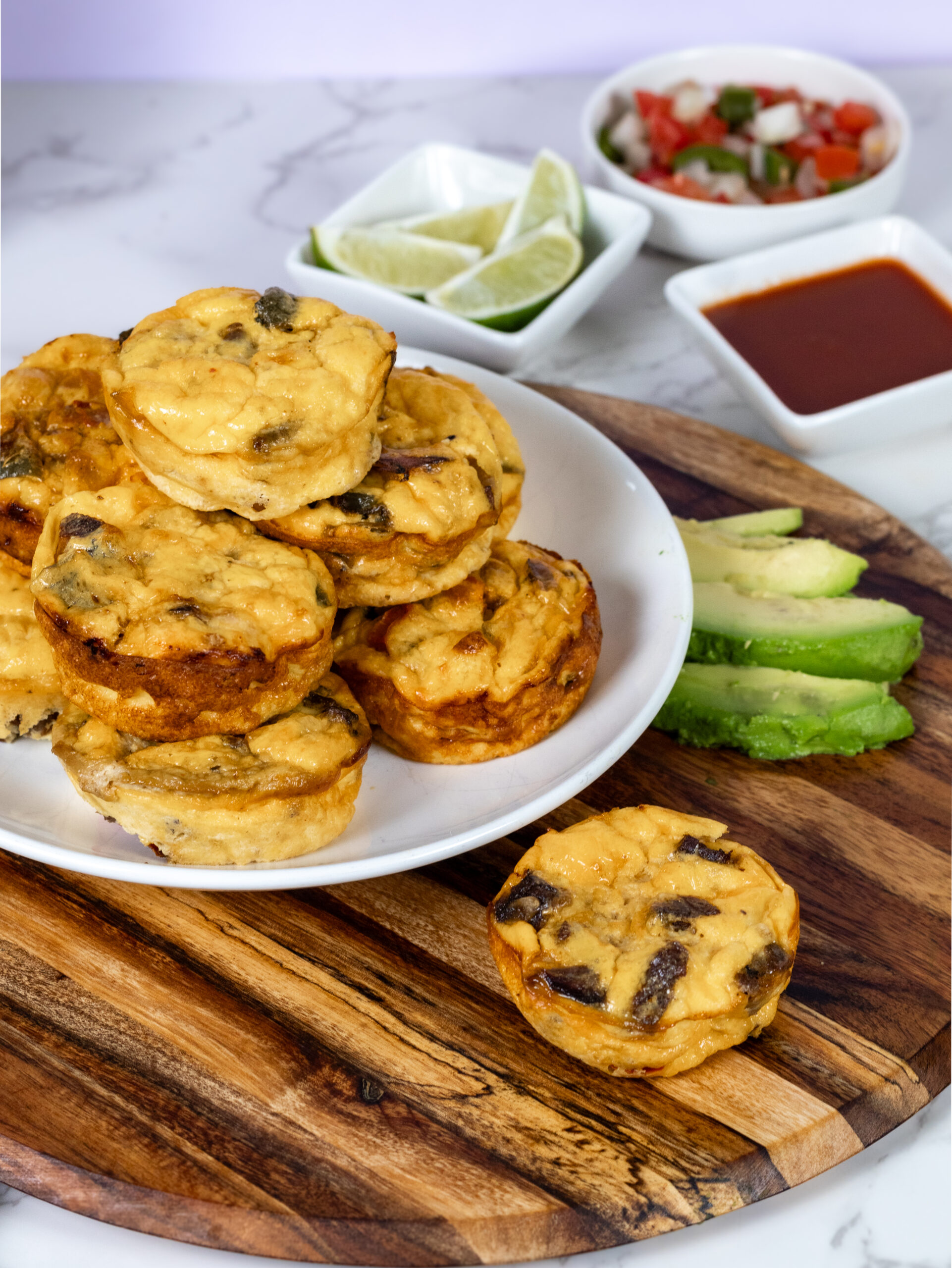 A plate stacked with savory muffins resembling fajita egg bites on a wooden board, accompanied by sliced avocados, lime wedges, and small bowls of sauce and salsa. The muffins are golden-brown with visible herbs. Marble surface and a soft purple background perfect for a high protein breakfast.