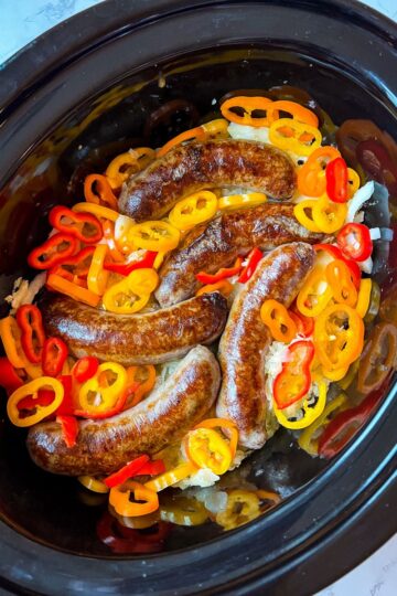 A slow cooker filled with browned beer brats topped with sliced red, orange, and yellow bell peppers. These ingredients are nestled on a bed of onions, creating a vibrant contrast in this brats and peppers crockpot meal.