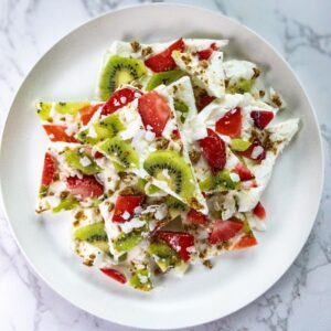 A plate of creamy Greek yogurt bark topped with kiwi slices, strawberry pieces, and crunchy granola. The bark is broken into irregular pieces and arranged on a white plate set against a marble background.