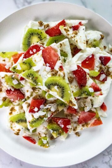 A plate of creamy Greek yogurt bark topped with kiwi slices, strawberry pieces, and crunchy granola. The bark is broken into irregular pieces and arranged on a white plate set against a marble background.