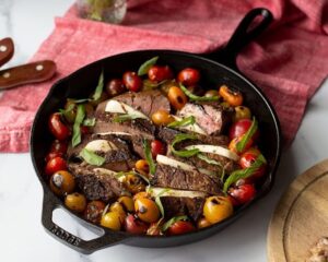 A cast iron skillet with sliced steak, mozzarella, cherry tomatoes, and fresh basil, resting on a marble surface. A red cloth and a wooden plate with a fork and knife are in the background.