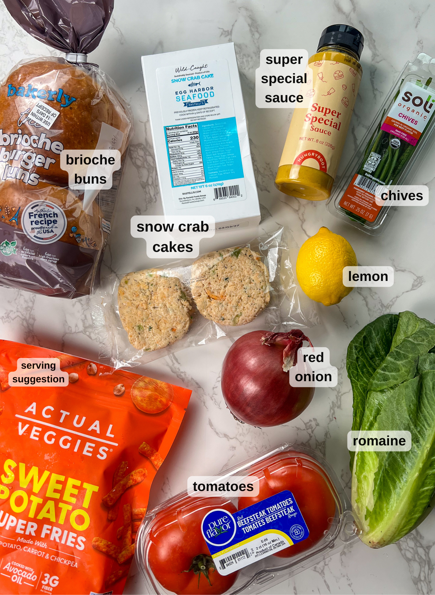 A variety of ingredients are arranged on a marble surface: brioche buns, snow crab cakes, super special sauce, chives, a lemon, a red onion, romaine lettuce, sweet potato fries, and tomatoes. Each item is labeled.