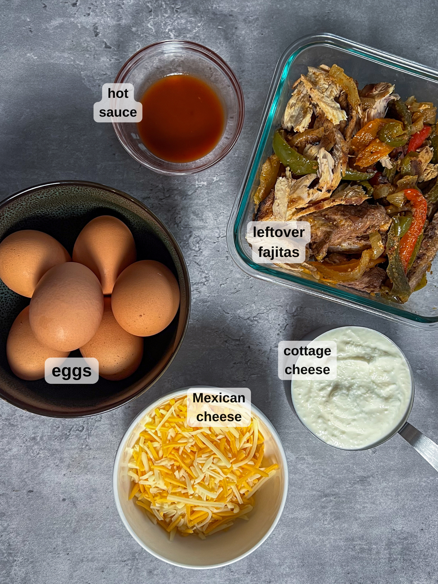 Top-down view of a kitchen counter with labeled items: a bowl of eggs for your breakfast egg bites recipe, a small dish of hot sauce, a container of leftover fajitas, a bowl of cottage cheese, and shredded Mexican cheese.