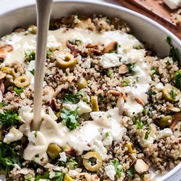 A bowl of crunchy quinoa salad with chopped green olives, parsley, nuts, and feta cheese is being dressed with a creamy sauce. In the background, a wooden cutting board holds chunks of feta cheese.