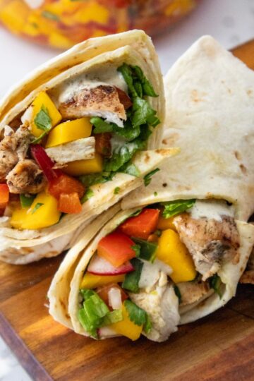 Two grilled chicken wraps with colorful fillings, including mango, red bell peppers, lettuce, and herbs, are neatly sliced and placed on a wooden board. A few pieces of grilled chicken are visible beside the wraps.