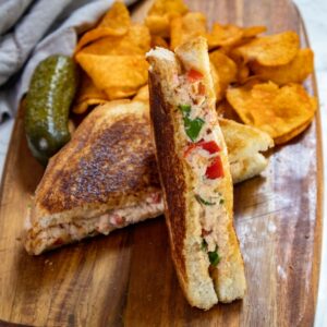 A grilled sandwich with visible filling of chicken, tomatoes, and spinach is cut in half and displayed on a wooden board. Accompanied by a pickle and a pile of potato chips in the background.