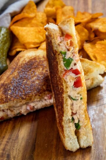 A grilled sandwich with visible filling of chicken, tomatoes, and spinach is cut in half and displayed on a wooden board. Accompanied by a pickle and a pile of potato chips in the background.