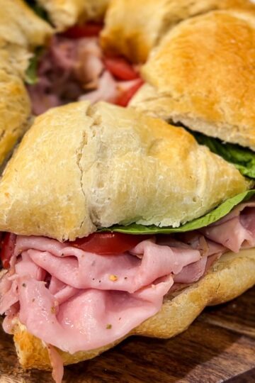 A circular sandwich with slices of ham, tomatoes, and lettuce, placed between thick, crusty bread. The sandwich rests on a wooden cutting board, and is sliced into individual portions.