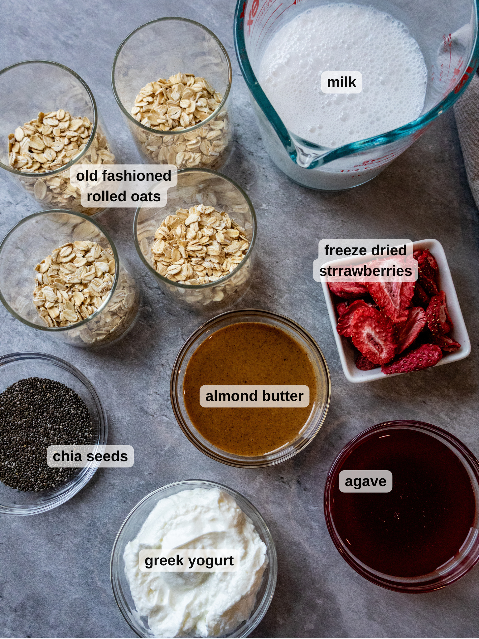 Ingredient spread for overnight strawberry overnight oats. 4 jars filled with oats, milk, strawberries, nut butter, chia seeds, yogurt, and agave syrup.