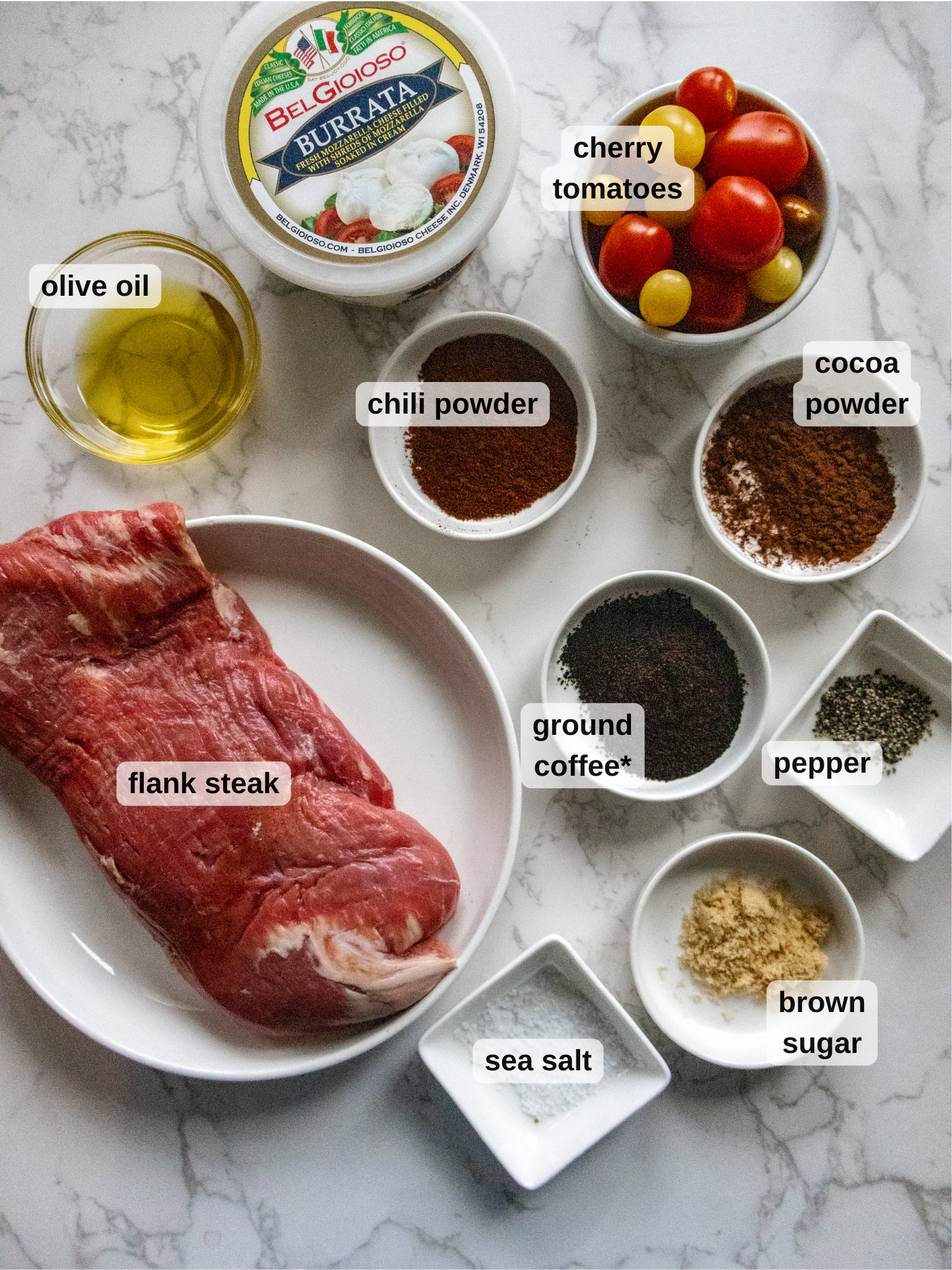 An array of ingredients on a marble surface showcases a raw flank steak ready for the perfect coffee rub. Surrounding it are small bowls of chili powder, cocoa, ground coffee, pepper, brown sugar, sea salt, olive oil, cherry tomatoes, and a container of burrata.