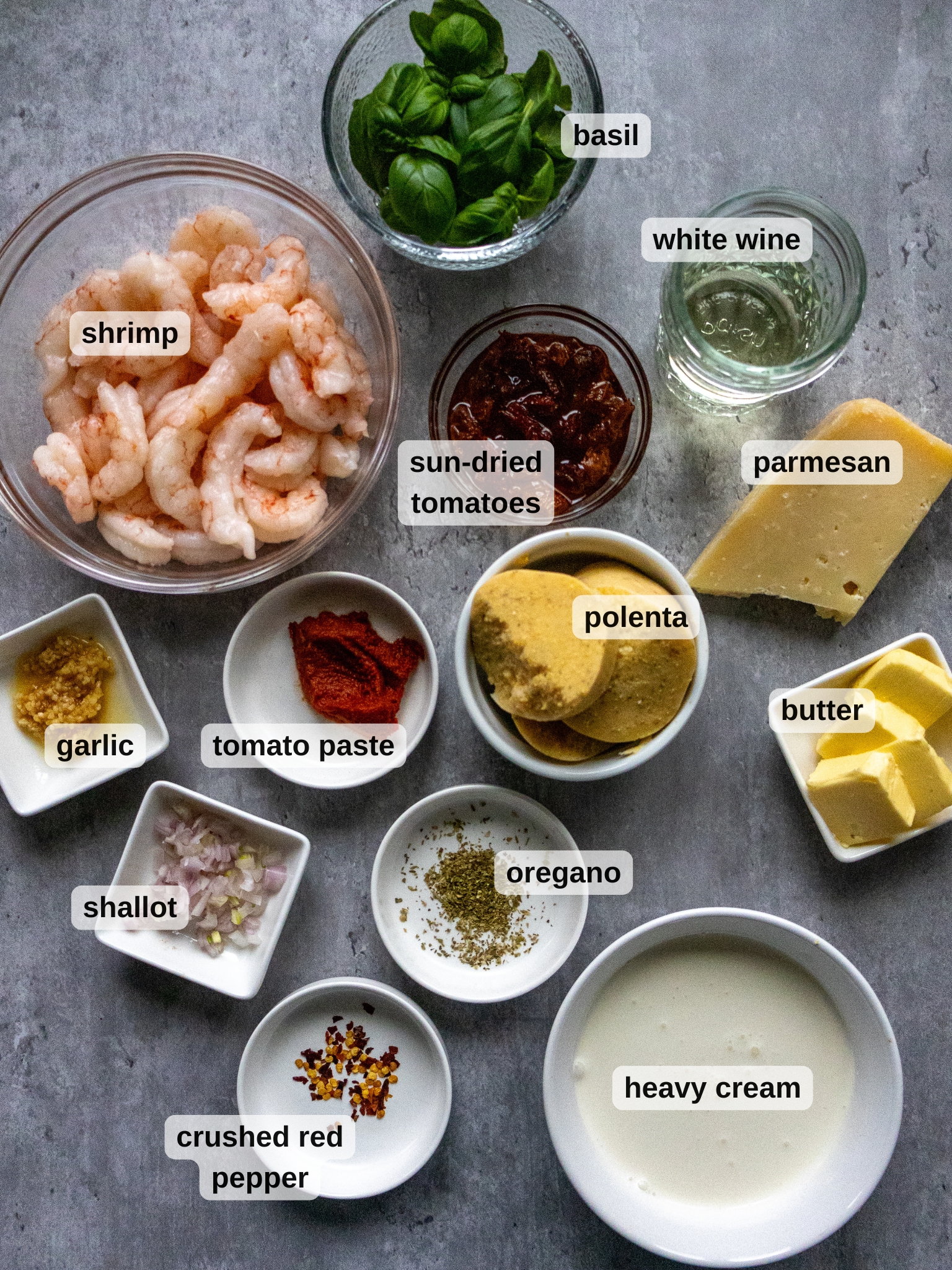 A perfect Valentines Day dinner spread awaits on the gray surface: a bowl of shrimp, fresh basil, white wine, sun-dried tomatoes, parmesan cheese, sliced polenta for a creamy parmesan polenta dish, butter, heavy cream, garlic, tomato paste, shallot, oregano and crushed red pepper.