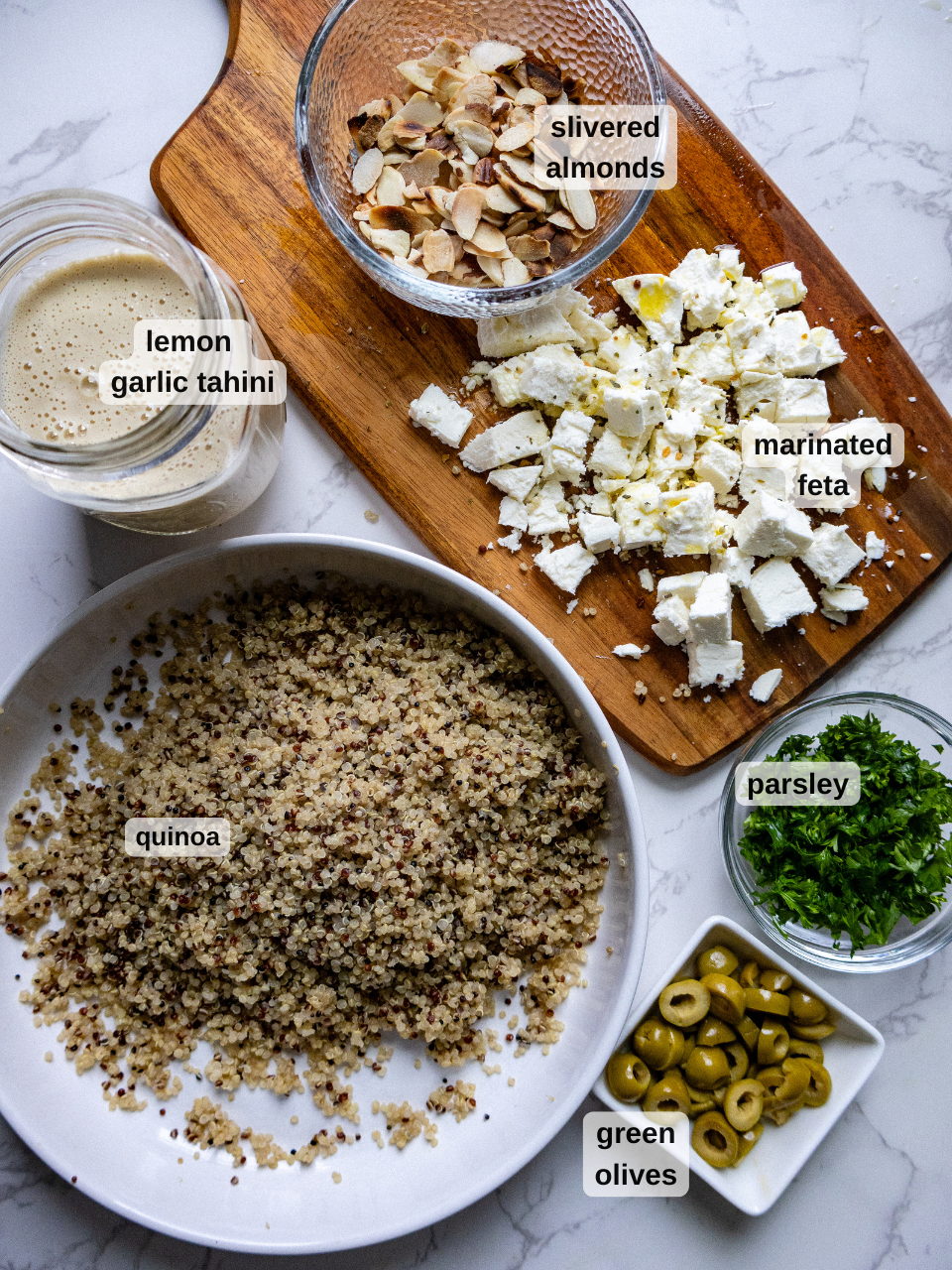 Top-down view of a kitchen counter with bowls of quinoa, chopped parsley, slivered almonds, marinated feta, lemon garlic tahini dressing, and green olives. The ingredients are labeled and neatly arranged for preparing a crunchy quinoa salad with green olives.