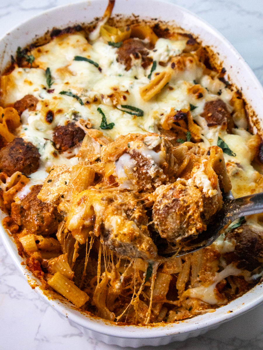 A baked pasta dish with meatballs, rigatoni, melted cheese, and herbs in a white casserole dish. A serving spoon lifts a portion, showcasing the stretchy, melted cheese. The dish is placed on a marbled surface.