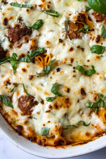 A baked rigatoni dish topped with melted cheese and fresh basil leaves, featuring frozen meatballs nestled among the pasta with golden-brown edges. Served in a white oval baking dish on a marble surface, this meal is both comforting and convenient.