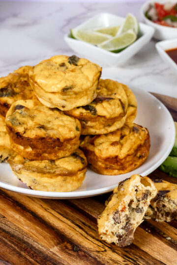 A plate of stacked mini breakfast casseroles with visible cheese and vegetables sits on a wooden board. These hearty fajita egg bites pair perfectly with sliced avocados, lime wedges, and small bowls of salsa, making for a delicious high protein breakfast.