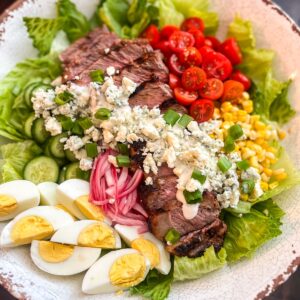 Enjoy a vibrant black and blue steak salad featuring sliced steak, cherry tomatoes, corn, blue cheese, cucumber, hard-boiled eggs, pickled onions, and lettuce. Its all topped with scallions and a drizzle of dressing for a perfect flavorful blend.