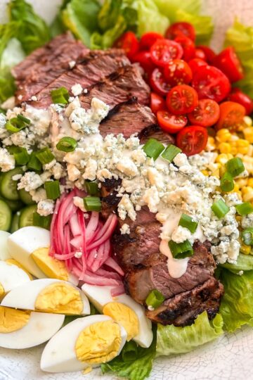 Enjoy a vibrant black and blue steak salad featuring sliced steak, cherry tomatoes, corn, blue cheese, cucumber, hard-boiled eggs, pickled onions, and lettuce. Its all topped with scallions and a drizzle of dressing for a perfect flavorful blend.