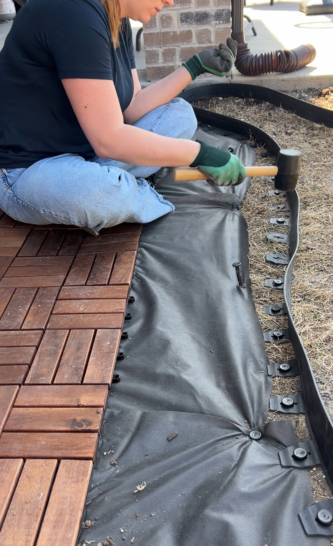 Person kneeling, wearing gloves, hammers black plastic border into dried grassy ground next to interlocking wooden deck tiles. A partially assembled area is visible, with various tools and materials around.