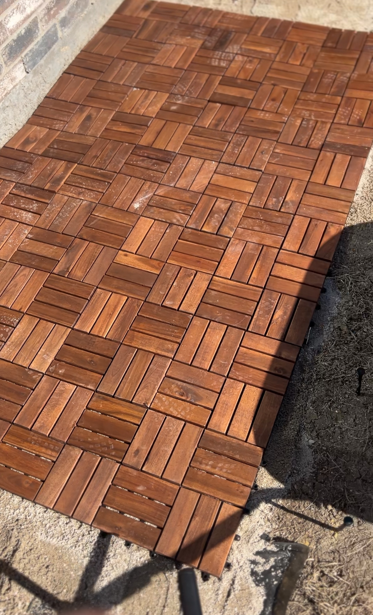 A wooden deck with interlocking tiles laid over dry ground. The tiles are arranged in a geometric pattern, showing varying shades of brown. Bright sunlight casts shadows on part of the deck.