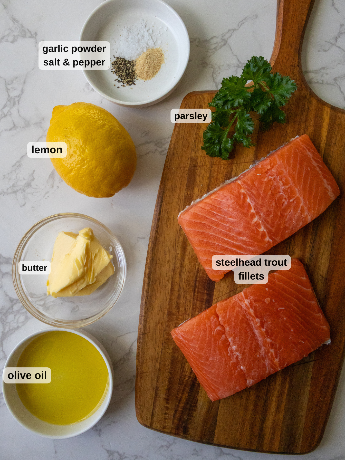 Two raw steelhead trout fillets on a wooden board with parsley. A lemon, garlic powder, salt, pepper, butter, and olive oil are nearby, each labeled. The background is a marble surface.