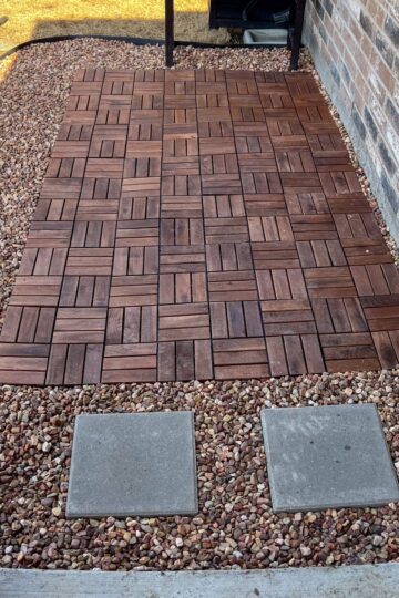 A small patio with interlocking wooden tiles is bordered by gravel. Two stone pavers lead to the patio from a concrete surface. A brick wall is on the right side of the patio area. Gardening tools are visible in the background.
