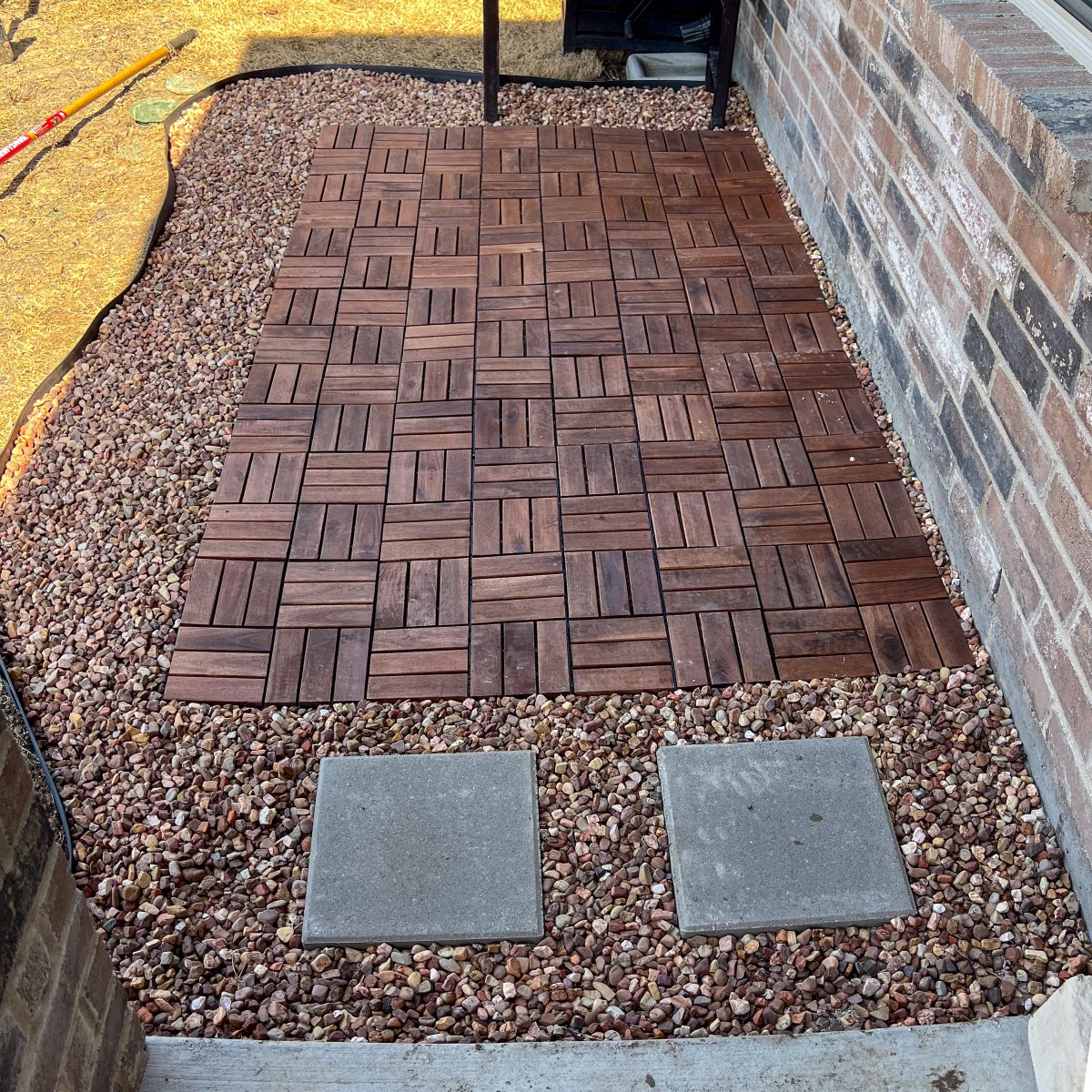 A small patio with interlocking wooden tiles is bordered by gravel. Two stone pavers lead to the patio from a concrete surface. A brick wall is on the right side of the patio area. Gardening tools are visible in the background.