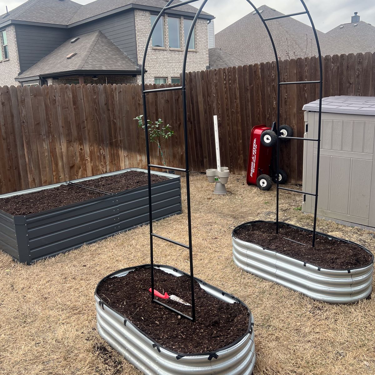 A backyard features two raised metal garden beds topped with trellises and filled with soil, ready for planting. A red toy wagon is propped vertically against a fence. A wooden fence and storage shed are visible in the background.