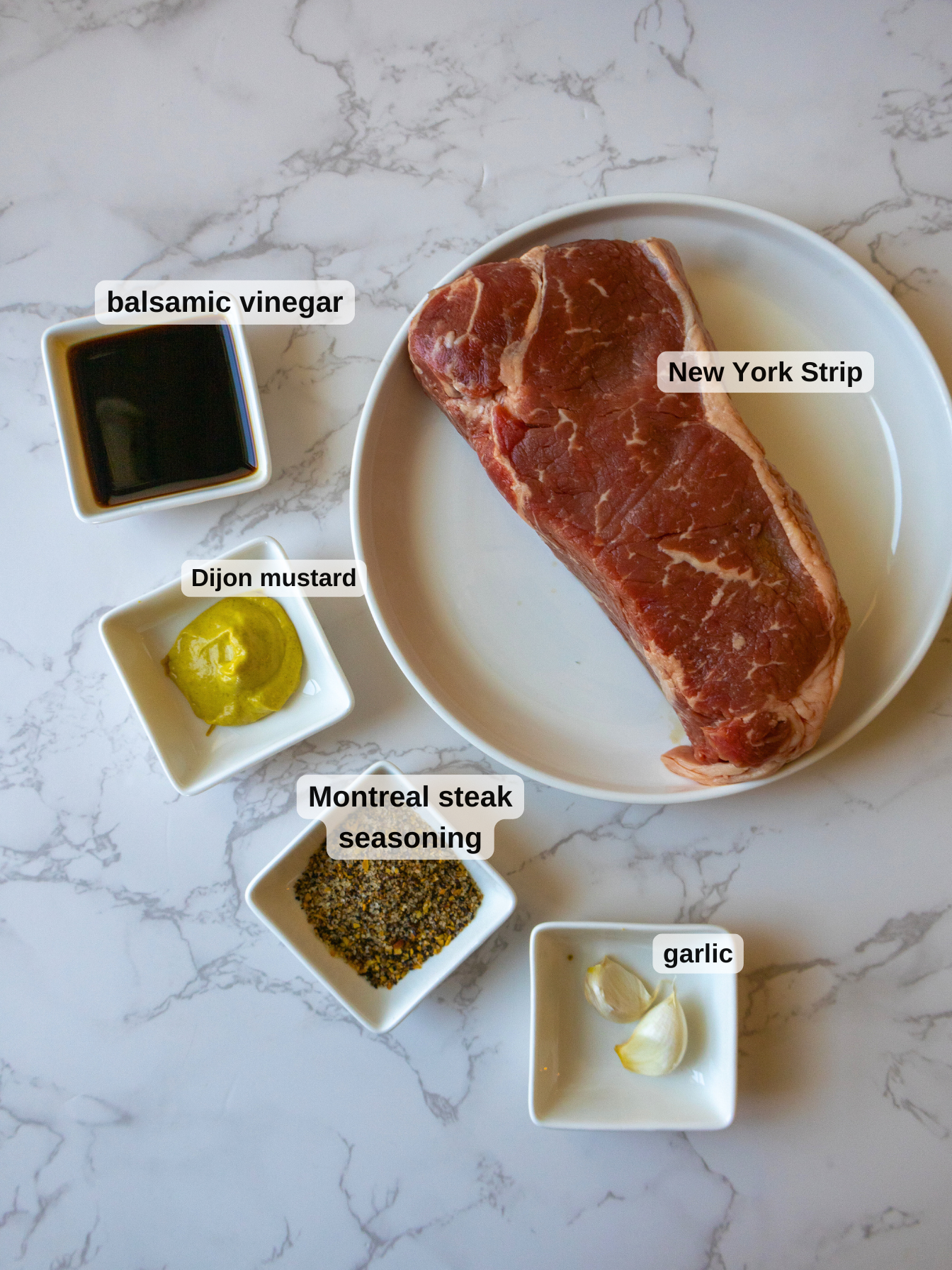 A raw New York strip steak sits on a white plate, accompanied by small bowls of balsamic vinegar, Dijon mustard, Montreal steak seasoning, and garlic cloves. This setup is perfect for crafting a flavorful balsamic steak salad on a marble surface.