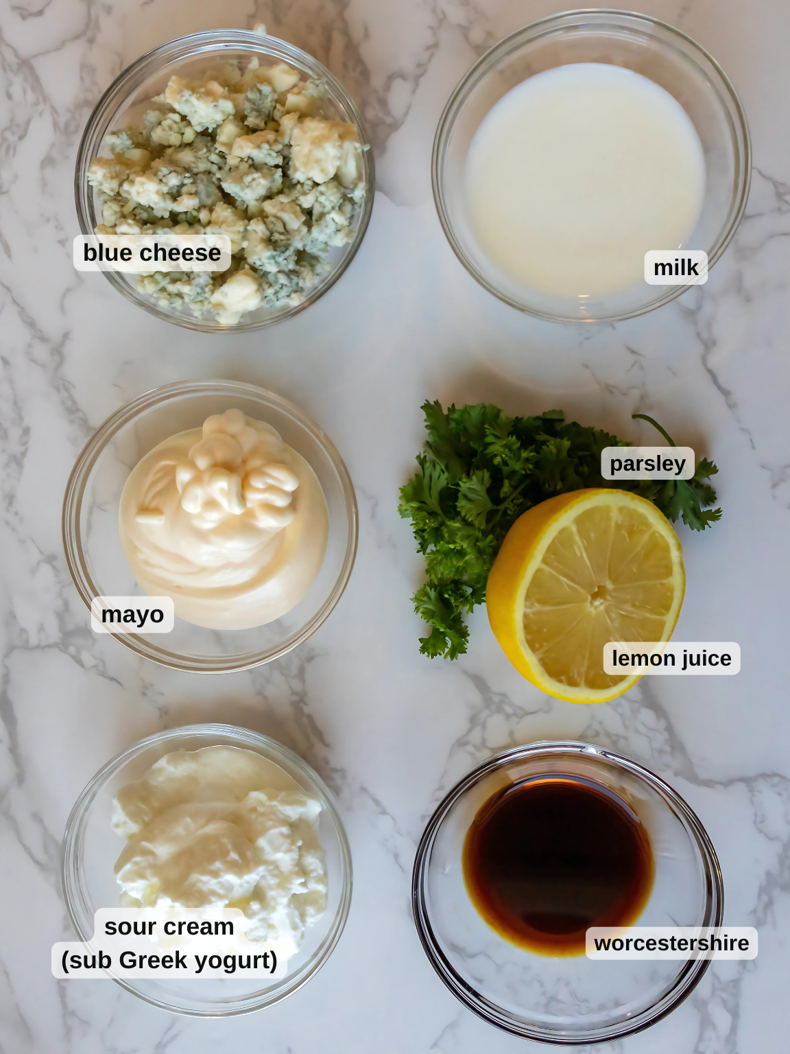Overhead view of six bowls on a marble surface, ready to whip up a black and blue salad. Ingredients include blue cheese, milk, mayo, sour cream (or Greek yogurt), Worcestershire sauce, parsley, and a half lemon for juice. Perfect for enhancing your steak and blue cheese salad experience.