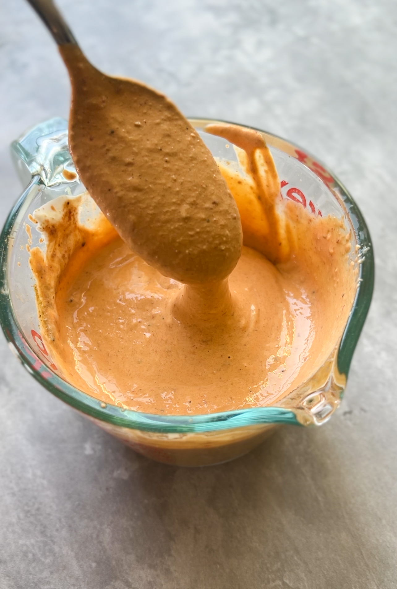 A spoon holds up a thick, creamy orange sauce above a glass measuring cup filled with the same sauce, set on a gray surface.