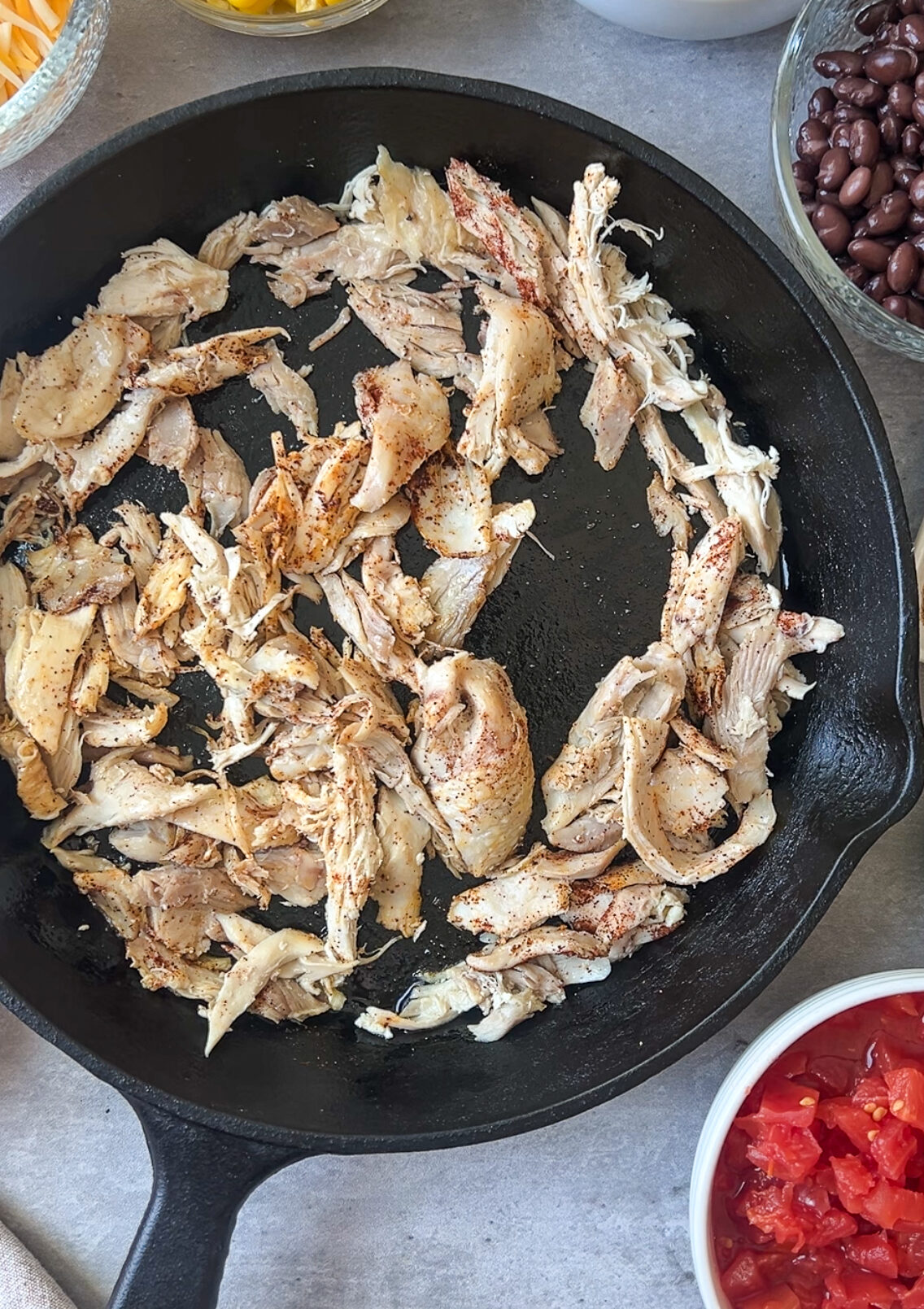 A skillet with shredded chicken surrounded by bowls of corn, chickpeas, black beans, shredded cheese, diced tomatoes, lime half, and seasoning powders on a gray surface.