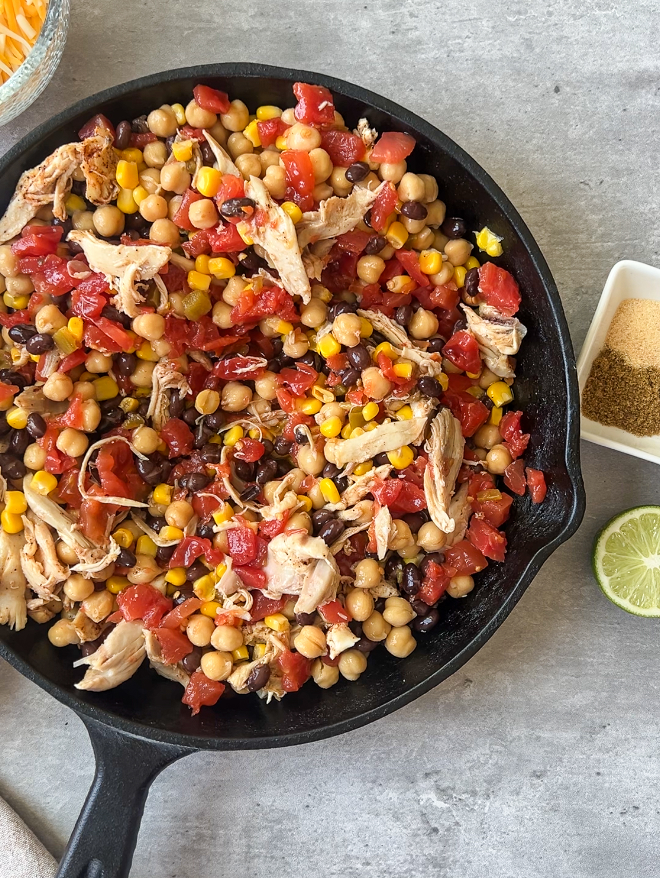 A skillet filled with a colorful mix of chickpeas, corn, black beans, diced tomatoes, and shredded chicken. It is garnished with lime zest. Next to the skillet is a halved lime and small bowls with spices.