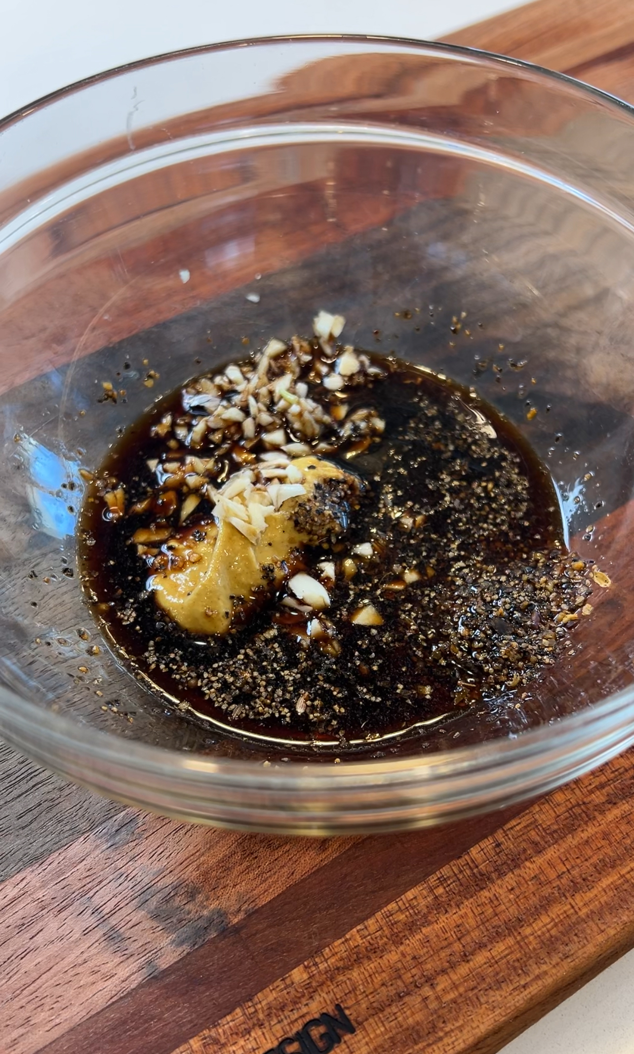 A glass bowl filled with a dark marinade mixture, including visible chunks of chopped garlic, black pepper, and a dollop of mustard, all combined in a soy sauce base. Perfect for enhancing the flavors of your favorite black and blue steak salad. The bowl is placed on a wooden surface.